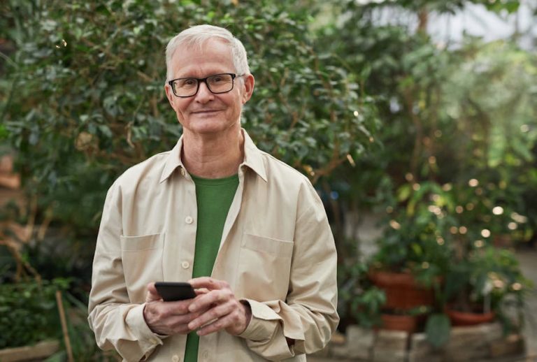 Senior man standing in a greenhouse garden, smiling while using a smartphone. Natural sunlight.